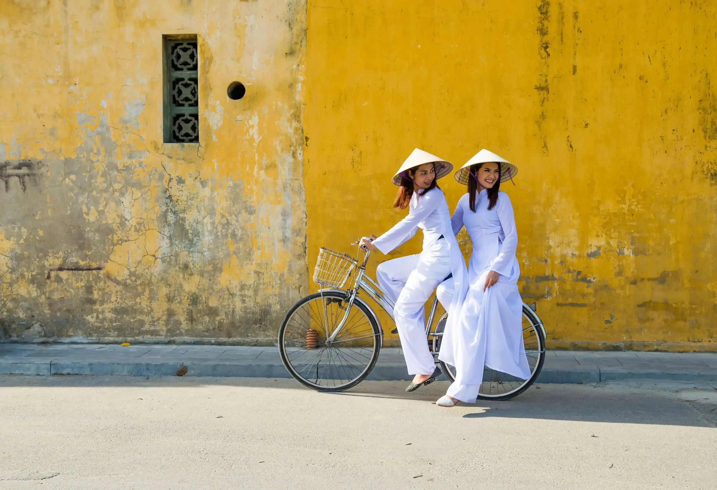Two women wearing white traditional clothes and conical hats riding on a bike along a dingy yellow-painted wall.