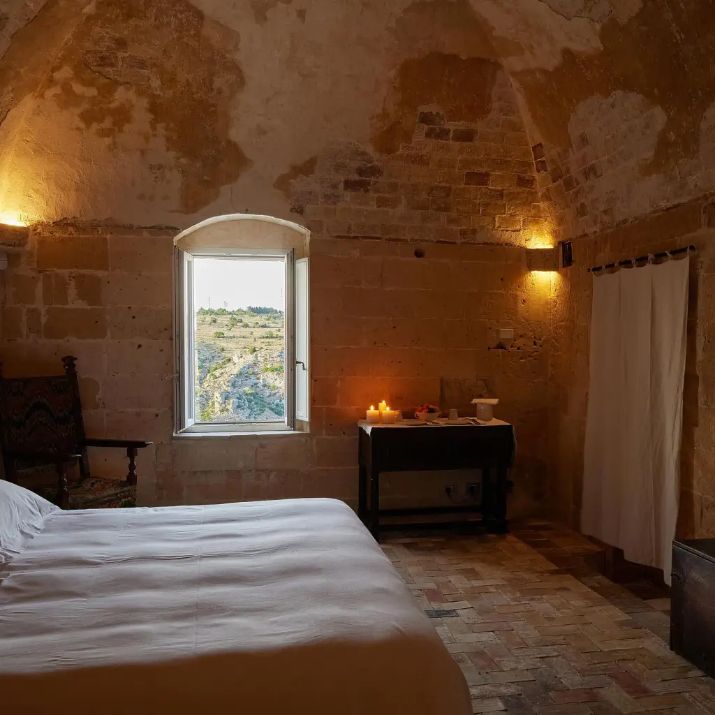 Cozy rustic bedroom with stone walls, a double bed, and small window showcasing countryside view. Warm glow from candles and wall lights creates a serene atmosphere.
