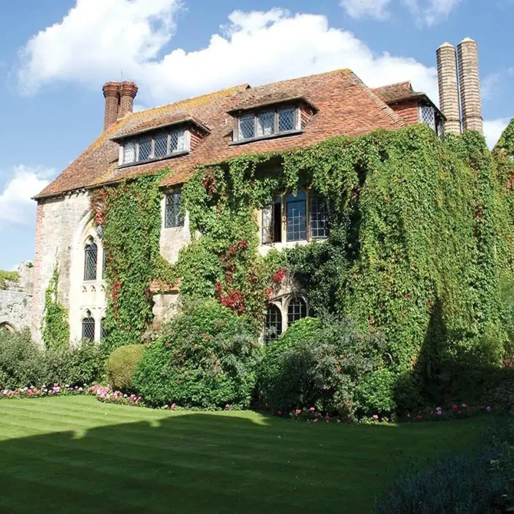Historic stone building with ivy-covered walls and arched windows, surrounded by lush gardens and manicured lawn under a clear blue sky.