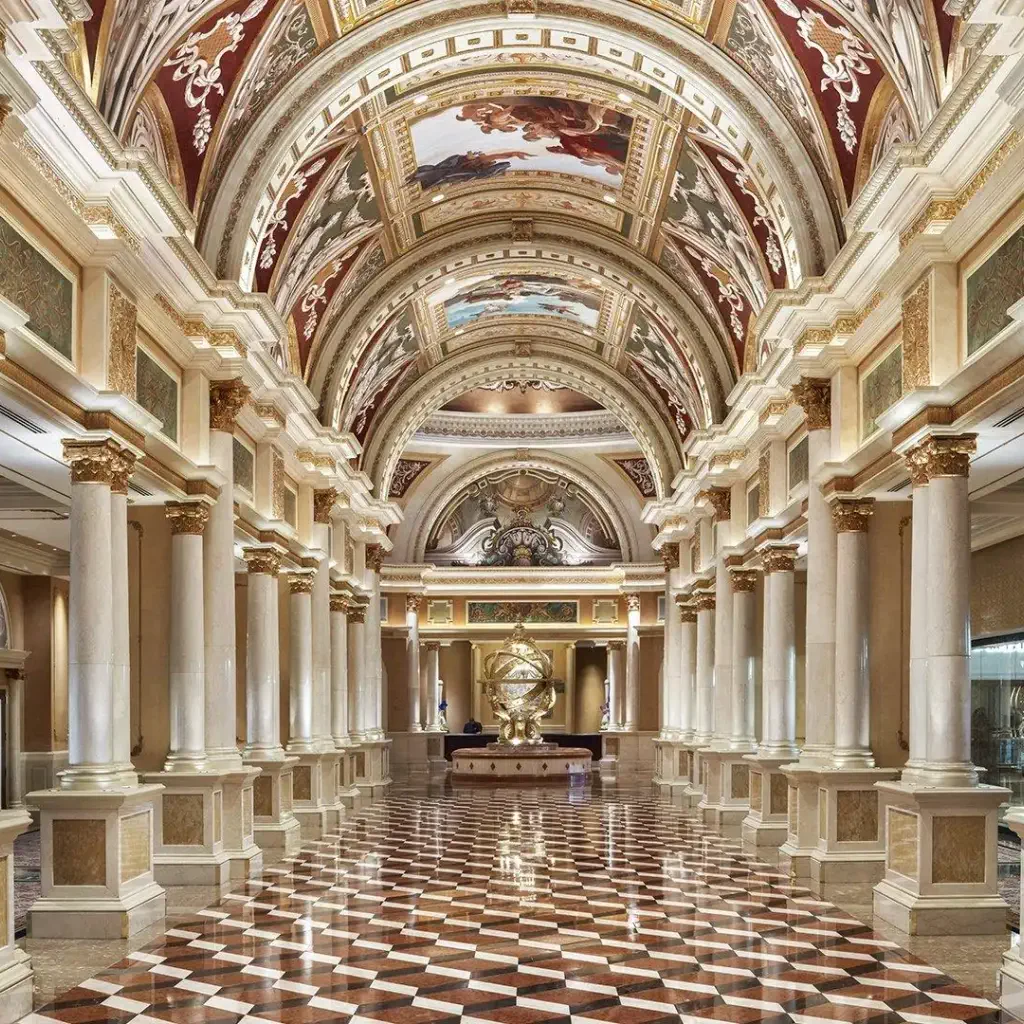 Ornate, high-ceiling corridor with marble columns, intricate frescoes, and a reflective checkered floor. A grand, opulent atmosphere is conveyed.