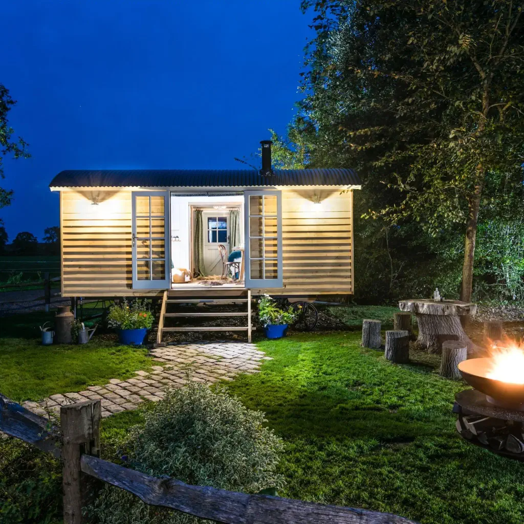 Cozy wooden cabin lit warmly at night, surrounded by lush greenery. A fire pit burns brightly on a lawn with outdoor seating, creating a tranquil atmosphere.