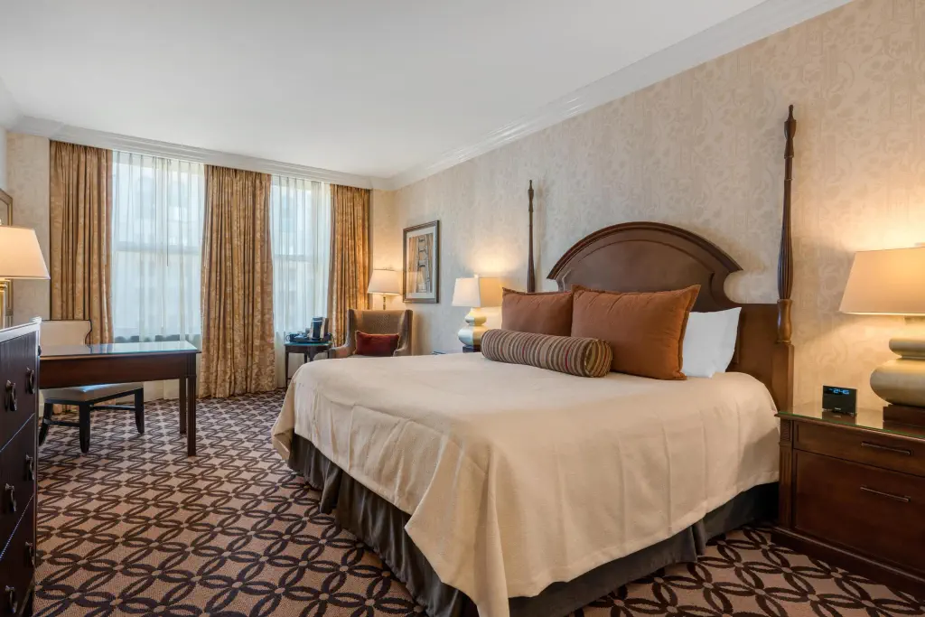 Hotel room with a king-sized bed, decorative cushions, and elegant furnishings. A desk, armchair, and lamps create a cozy, inviting atmosphere.