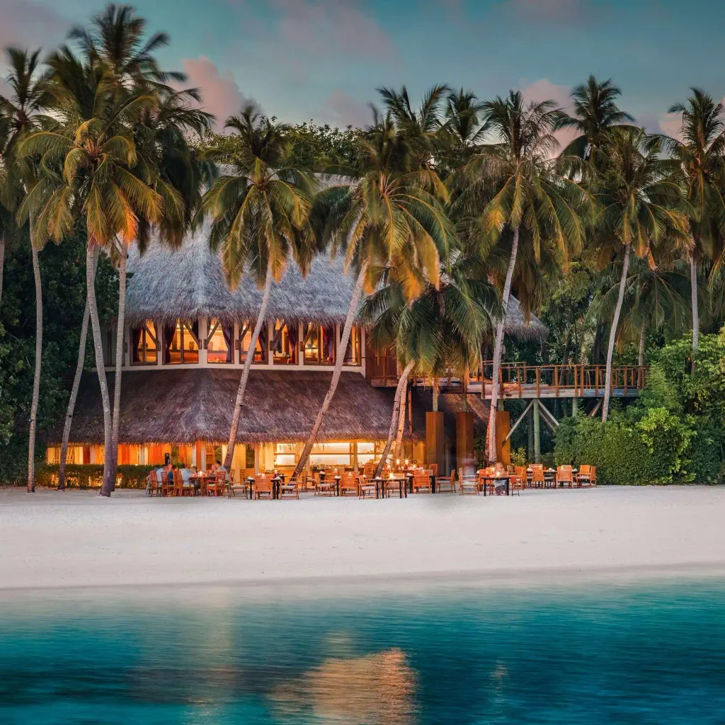 Tropical beach scene at dusk with a straw-roofed resort surrounded by palm trees. Warm lights glow from the building, reflecting on calm, turquoise water.