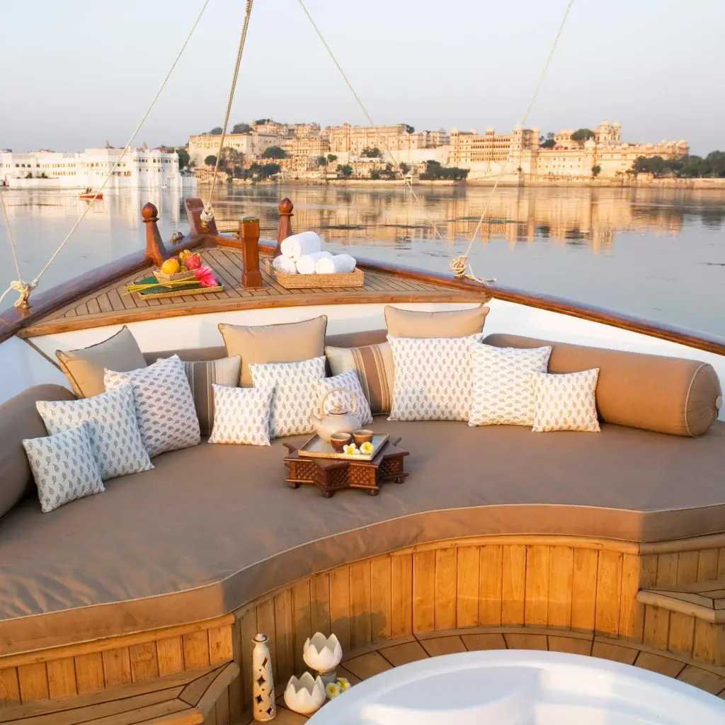 Luxurious boat deck with cushioned seating, pillows, and a small table with fruit and drinks. Scenic view of a serene lake and palace at sunset.