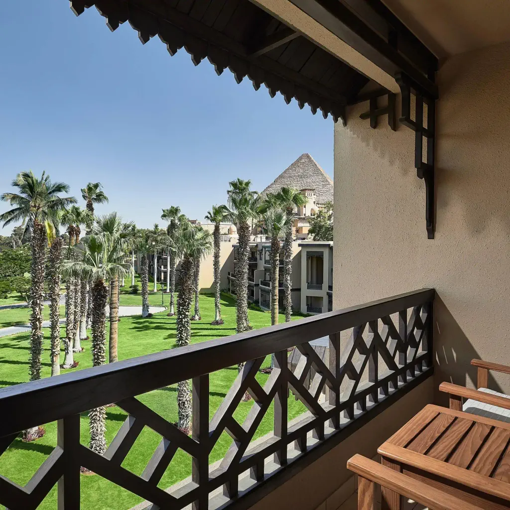 Balcony view with two chairs overlooking a lush garden filled with tall palm trees. In the background, a pyramid is visible under a clear blue sky.