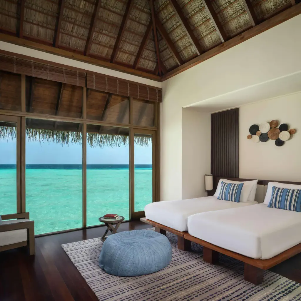 Luxurious oceanfront room with twin beds, wooden decor, large windows offering a stunning turquoise sea view. Calm, serene, and inviting atmosphere.