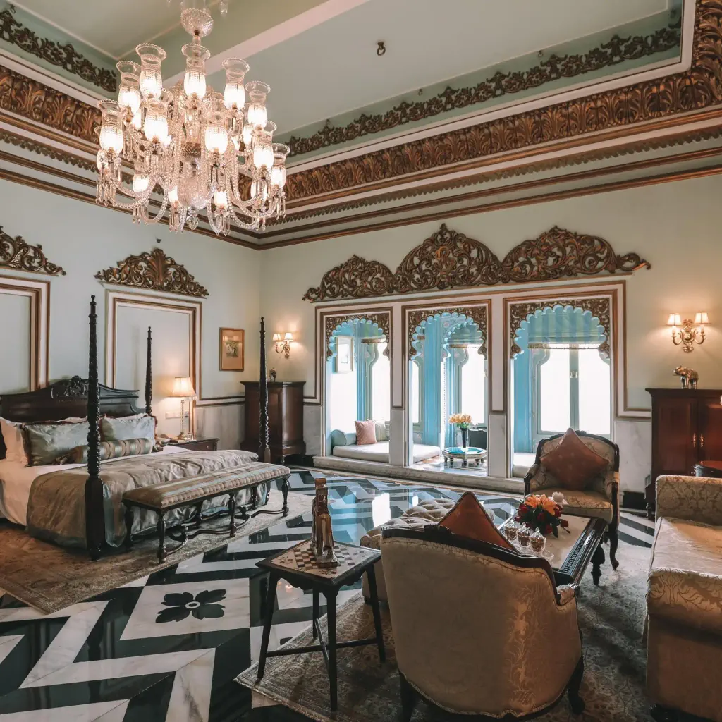 Luxurious bedroom with ornate ceilings, elegant chandelier, and opulent decor. Features a large bed, sofas, and windows with blue accents.