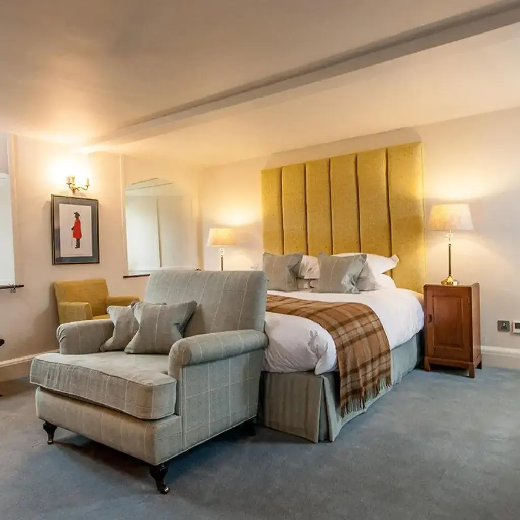 Elegant hotel room with soft lighting. Features a plush bed with a yellow headboard, cozy armchair, wall art, and a view into a modern bathroom.