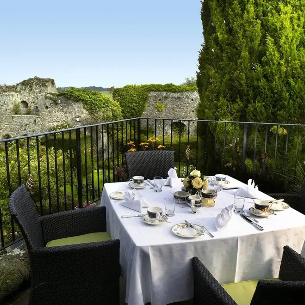 Outdoor dining on a terrace with a white tablecloth, tea set, and flower centerpiece. Surrounded by lush greenery and stone ruins in the background.