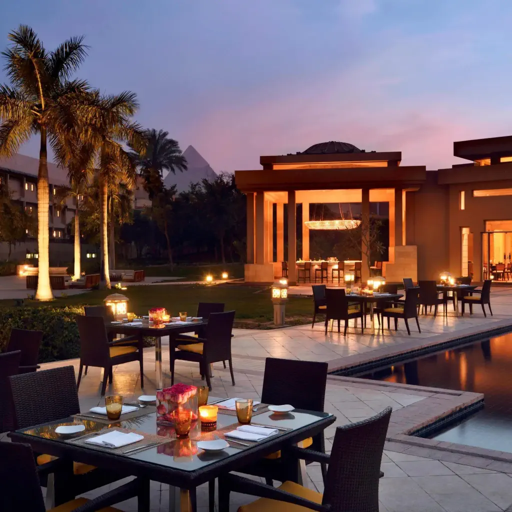 Outdoor dining area at sunset with tables set for dinner by a lit pool, surrounded by palm trees. Warm, inviting ambiance with soft lights.