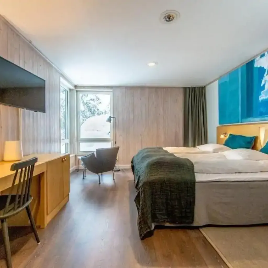 A cozy hotel room with wood paneling, a large bed, teal bedding, a desk with a lamp, a modern chair by a window, and an abstract blue painting.
