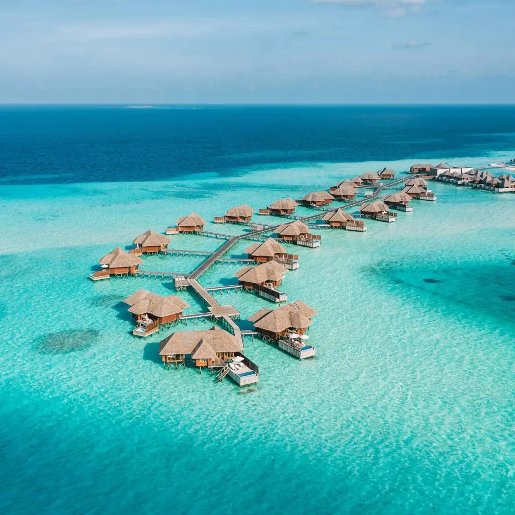 Aerial view of overwater bungalows on turquoise waters, connected by wooden walkways, set against a backdrop of deep blue ocean and clear sky.