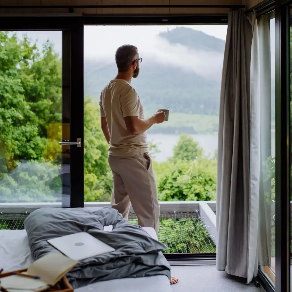 A man holds a cup of coffee as he stands beside a big glass window with vast nature views.