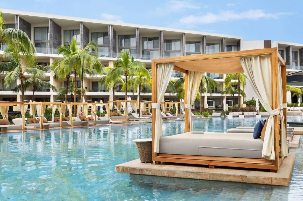 Poolside cabanas and lounge beds are arranged around a large swimming pool with palm trees, in front of a modern multi-story resort building.