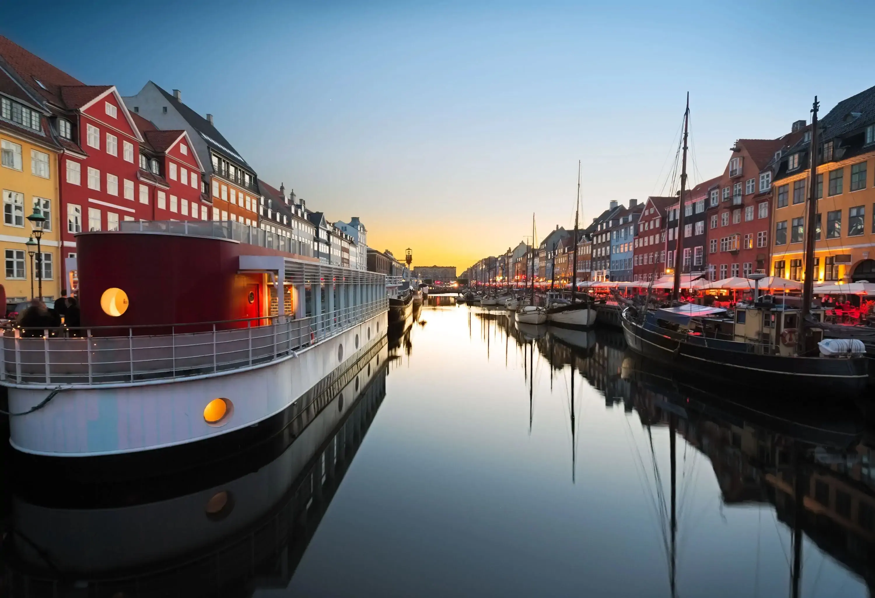 A tranquil canal lined with houseboats, sailboats, and colourful buildings during sunset.