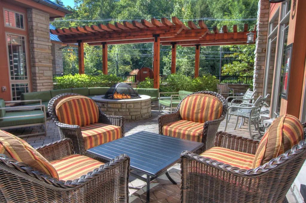 Outdoor patio with wicker chairs and striped cushions around a black table, a stone fire pit, and a wooden pergola, surrounded by greenery.
