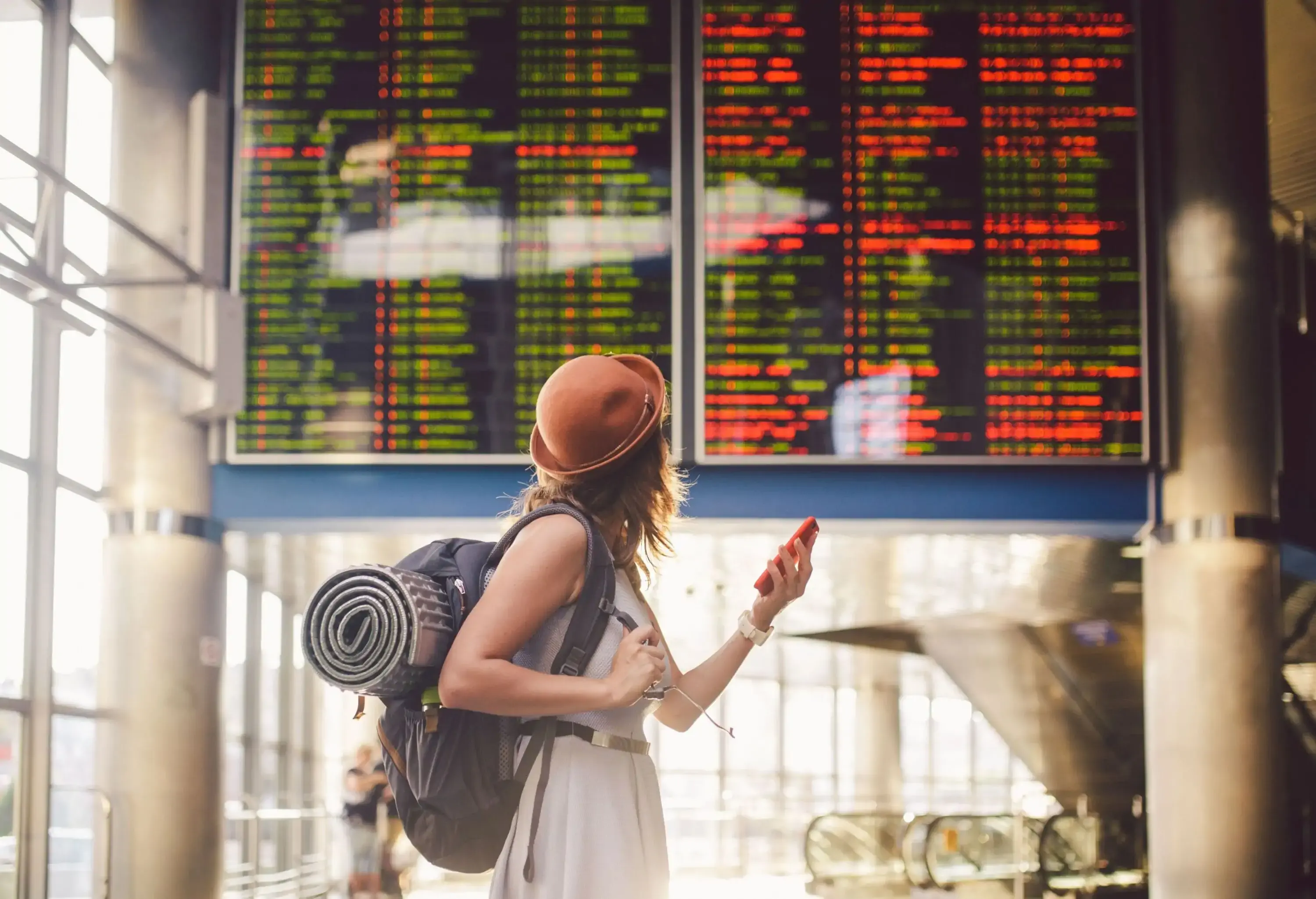 A female with a backpack holding a red phone as she looks at the electronic schedule board.