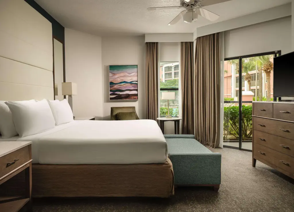 Hotel room with a large, white bed, wooden furniture, and a modern abstract painting on the wall. Large windows and curtains overlook greenery