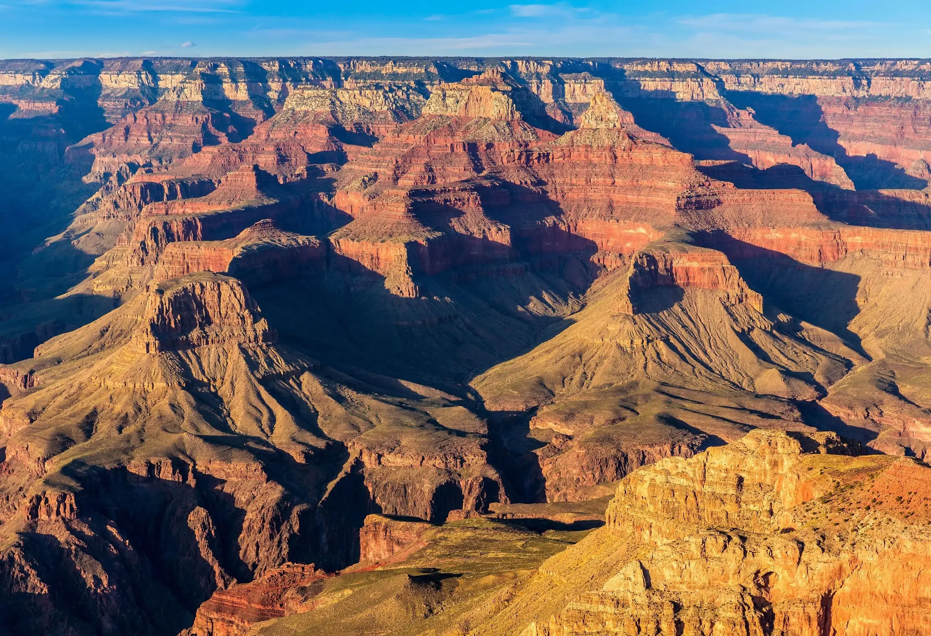 The sunlit Grand Canyon features red rock layers topped with an immense flat area of land.
