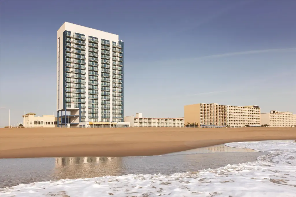 Beachfront high-rise hotel with glass balconies, adjacent low-rise buildings, and sandy beach in the foreground; calm ocean waves under a clear sky.