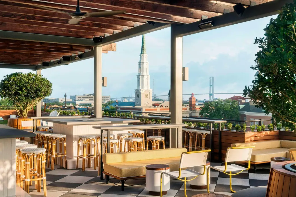 Rooftop bar with wooden ceiling, wicker stools, yellow sofas, and lush plants. Cityscape view features a church spire and distant bridge.