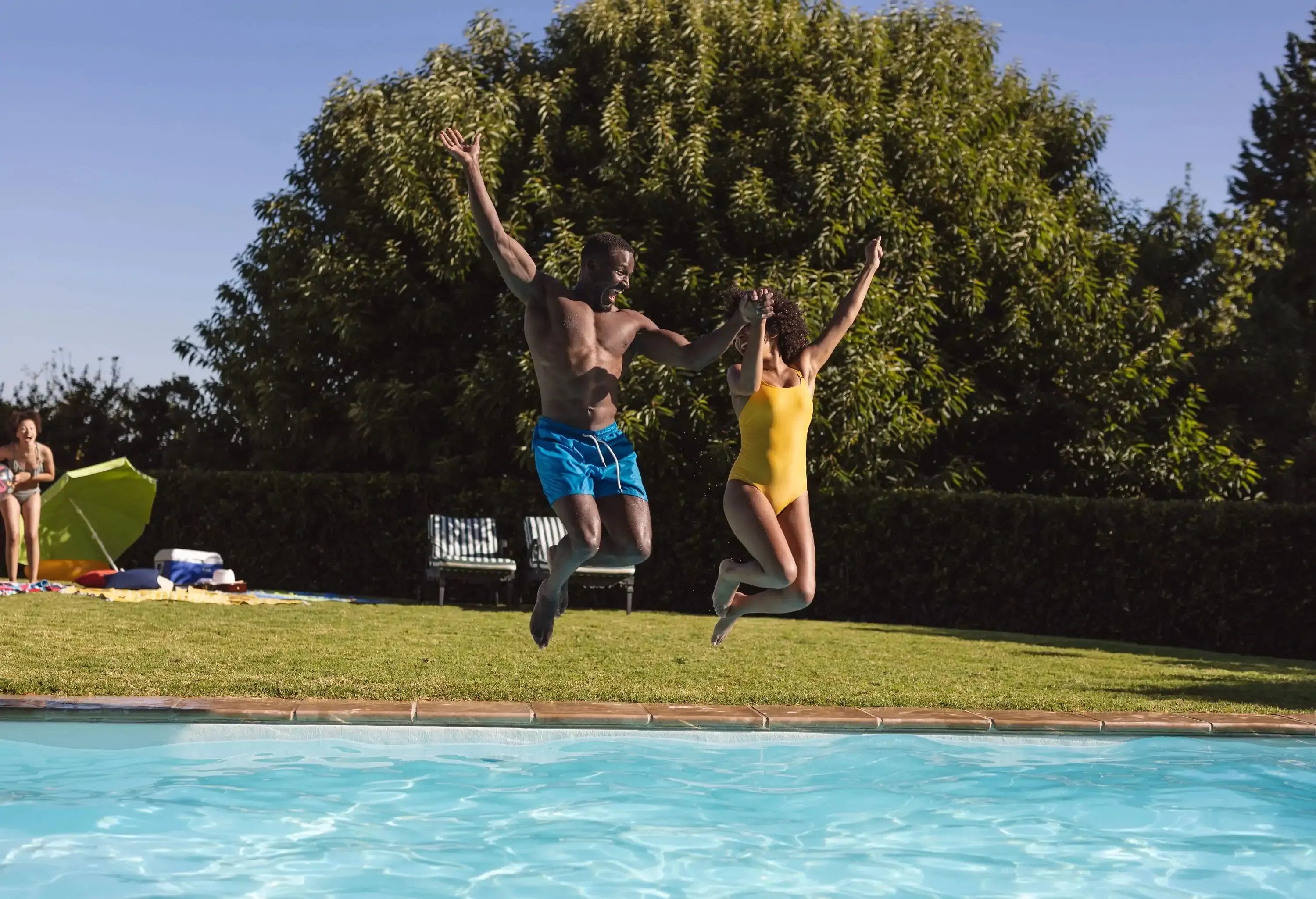 A couple wearing vibrant swimwear jump mid-air with arms spread out above a pool.