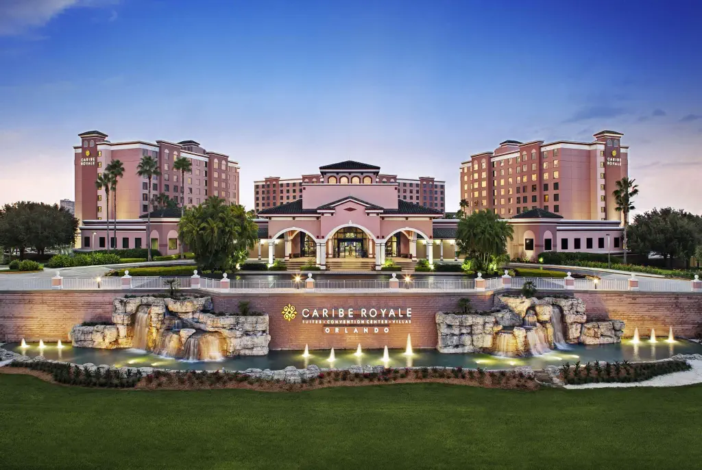Elegant resort exterior at sunset, showing a grand entrance with arches and palm trees, surrounded by illuminated fountains and a manicured lawn.