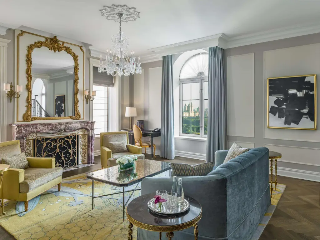 Luxurious living room with ornate fireplace and large mirror, crystal chandelier, plush sofas, abstract art, and a window with cityscape view. 