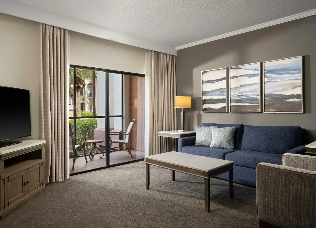 Modern living room with dark blue sofa, abstract art on gray wall, and a TV. Large window with open curtains reveals a patio with chairs.