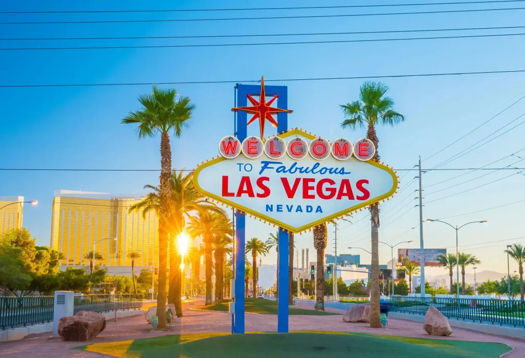 The well-known Las Vegas roadside sign, with sunlight beaming through the trunks of palm trees in the backdrop.