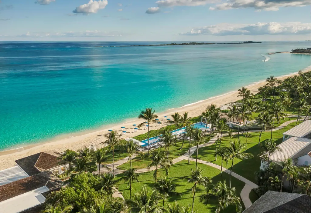 Tropical beachfront view featuring palm trees, a pristine sandy shore, turquoise waters, and a luxurious pool area in the foreground.