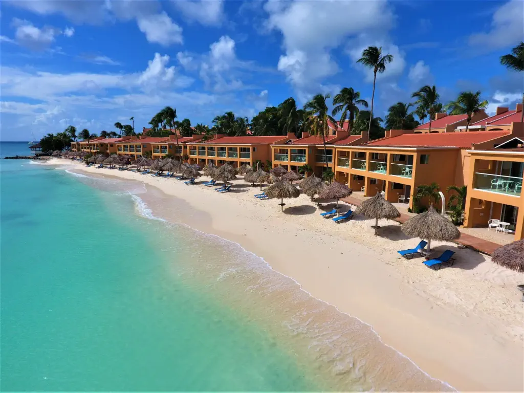 Coastal resort view with beachfront villas along a turquoise sea. Sandy beach lined with straw umbrellas, blue sun loungers, and lush palm trees under a clear sky.
