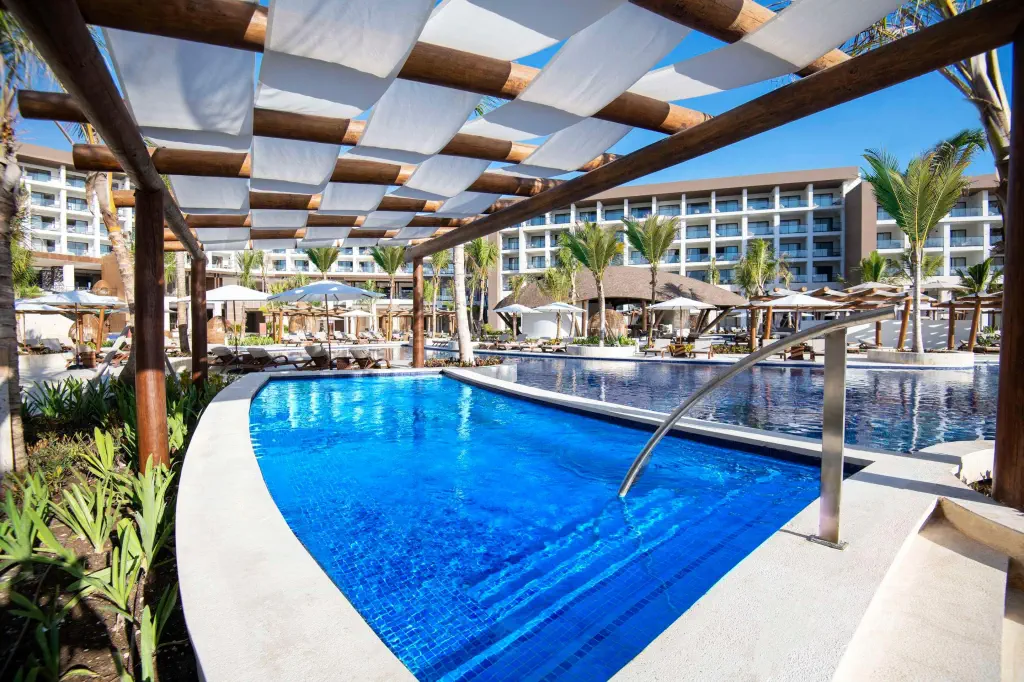 Luxurious resort pool area with clear blue water, surrounded by sun loungers, palm trees, a pergola, and a multi-story hotel building under a bright sky.