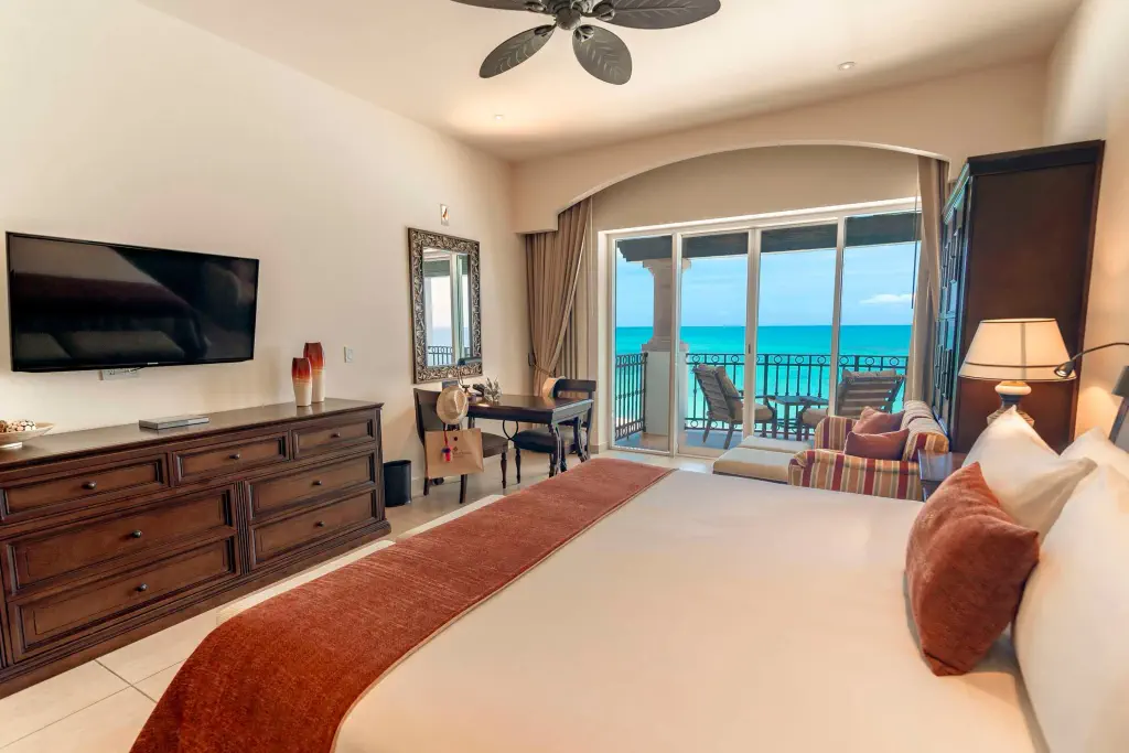A stylish hotel room featuring a large bed, wooden dresser, and balcony with ocean views, evoking a serene atmosphere.