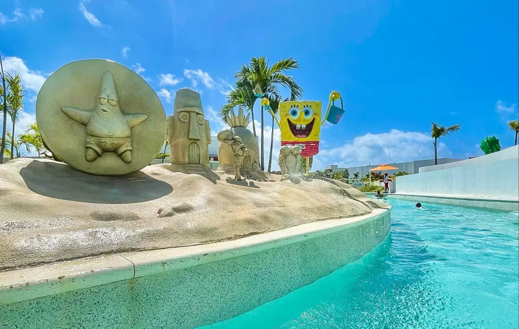 SpongeBob and friends' statues in a sandy beach scene near a turquoise pool. Lush palm trees, vivid blue sky. Joyful, sunny atmosphere.