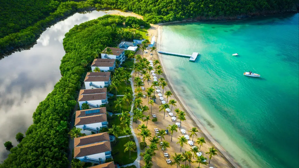 Aerial view of a tropical beach resort with villas along a lush coastline. Palm trees line the sandy beach, leading to turquoise water with anchored boats.