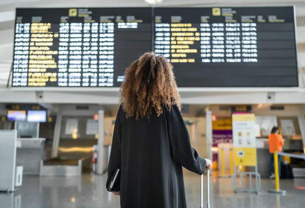 A curly-haired person in a black dress stands in front of an airport flight information display.