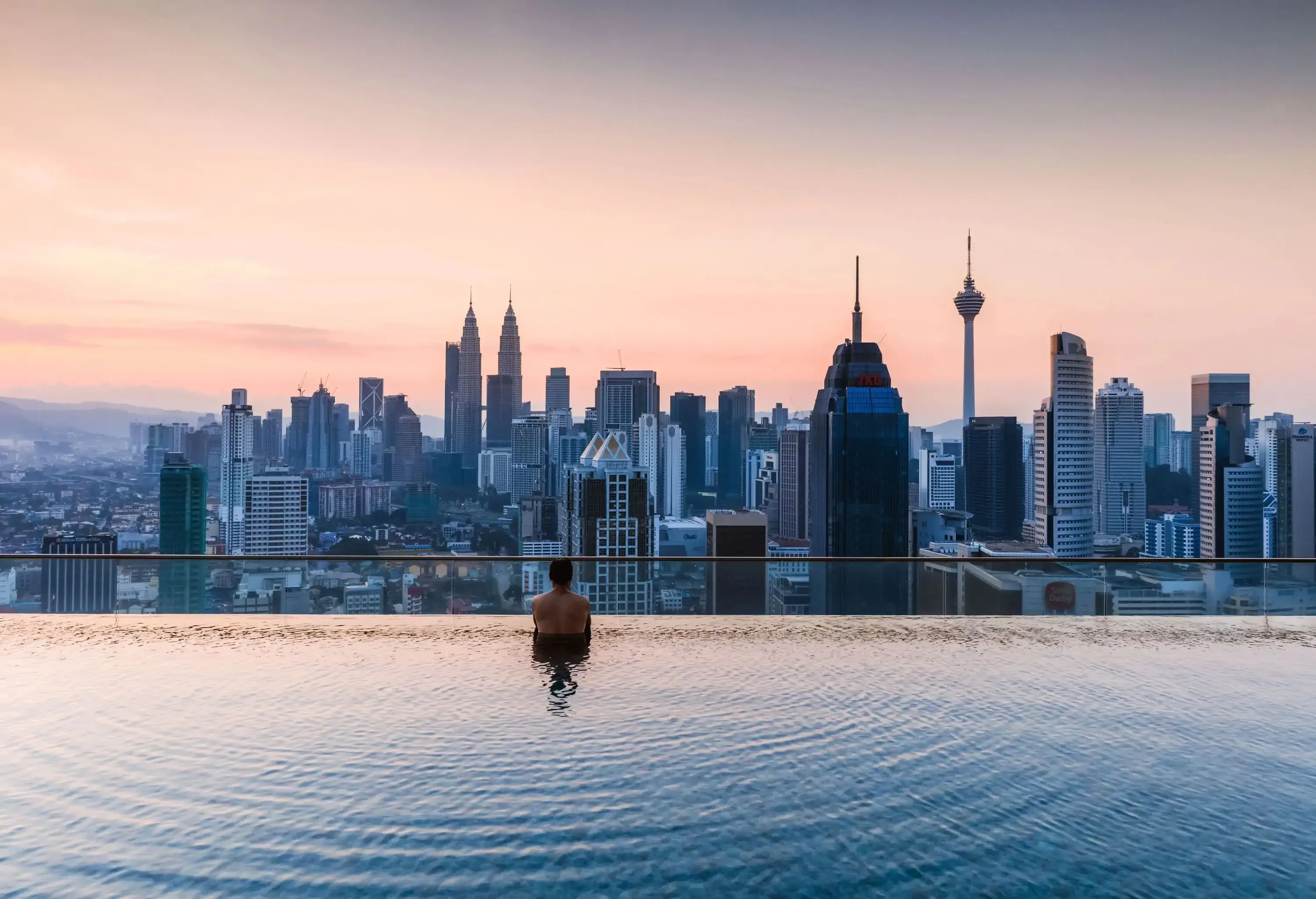 A man partially submerged on a rooftop infinity pool, looking at the beautiful skyline against the scenic pinkish sky.