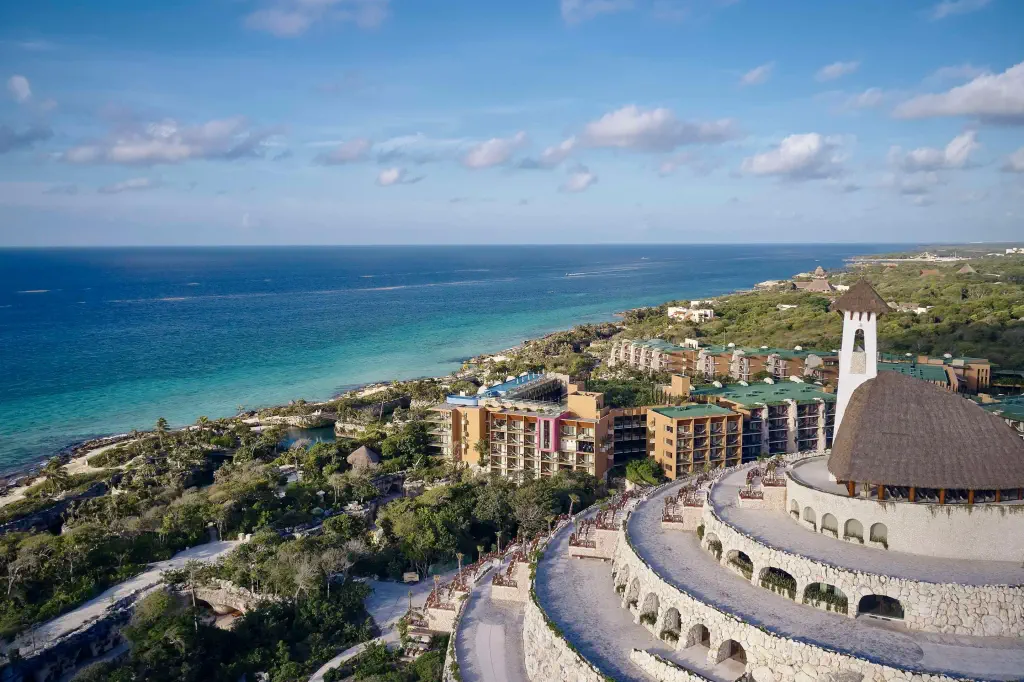 Aerial view of a seaside resort with lush greenery, colorful buildings, and turquoise waters under a clear blue sky.