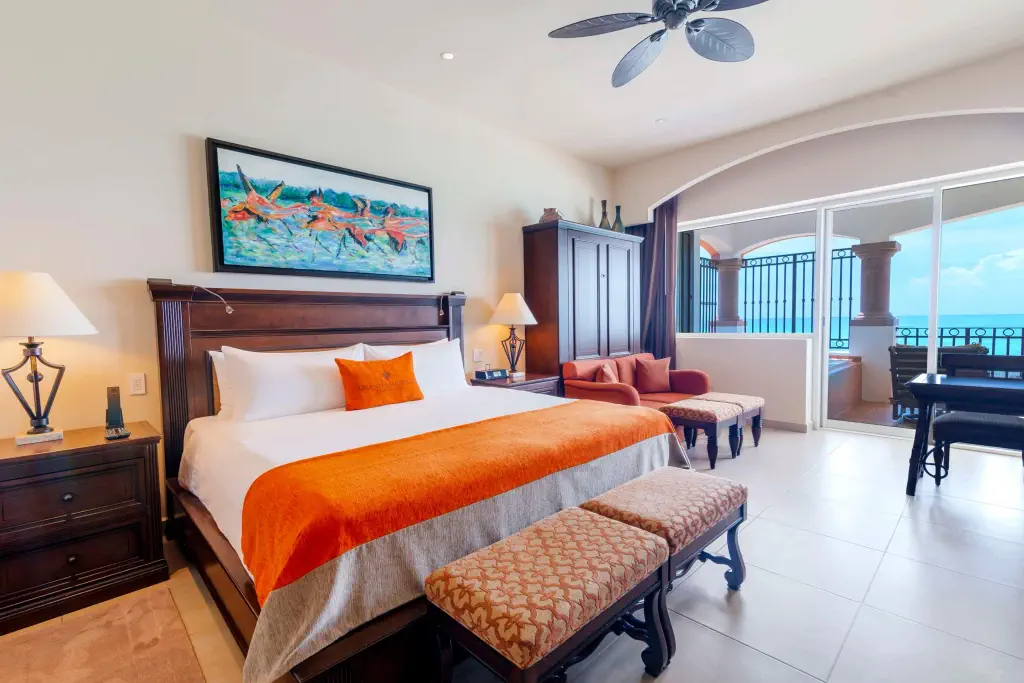 Bright and spacious hotel room featuring a king bed with orange accents, seating area, and large windows with ocean views.
