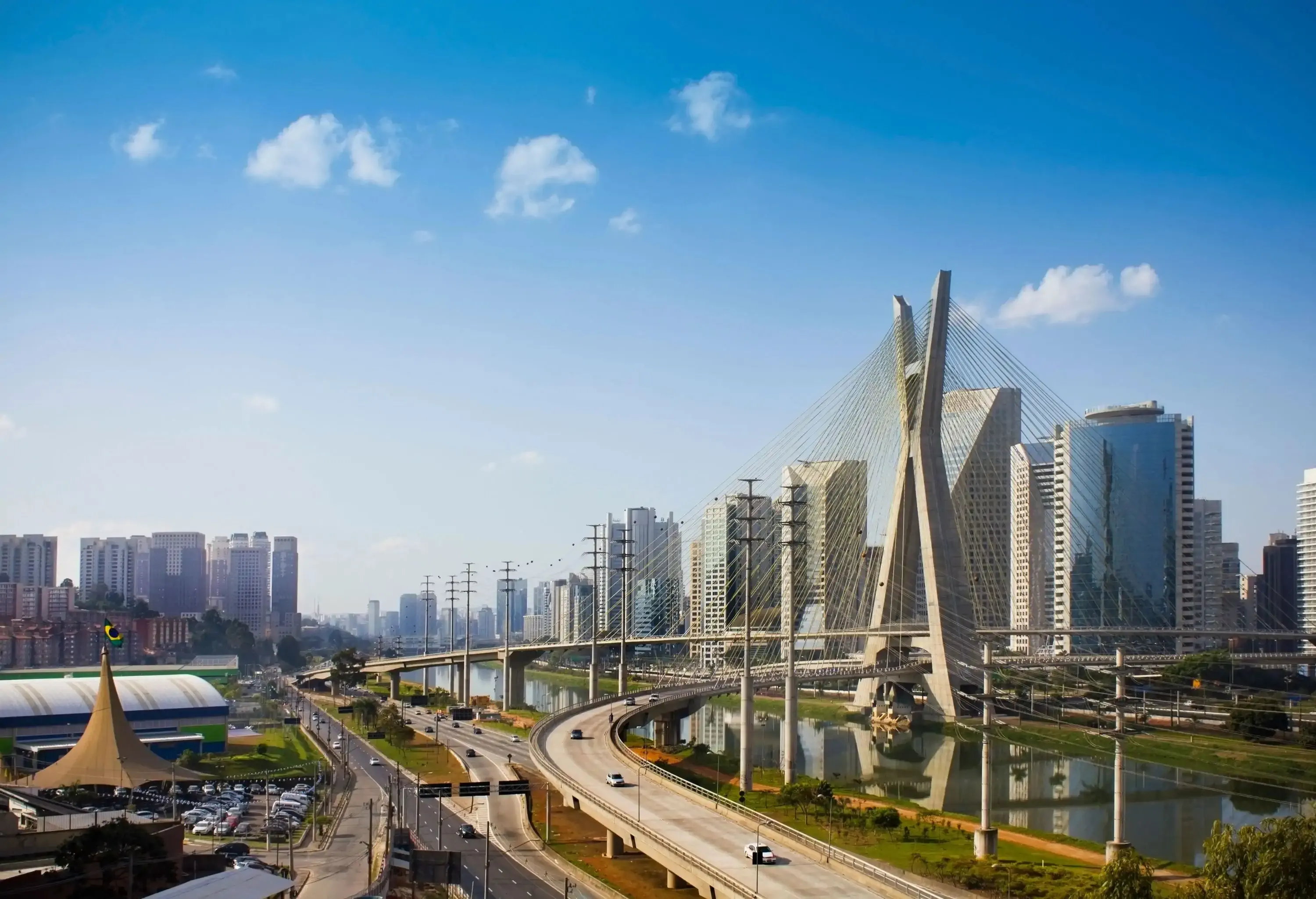 A cable-stayed bridge across a calm river bordered with roads and a cluster of high-rise buildings.