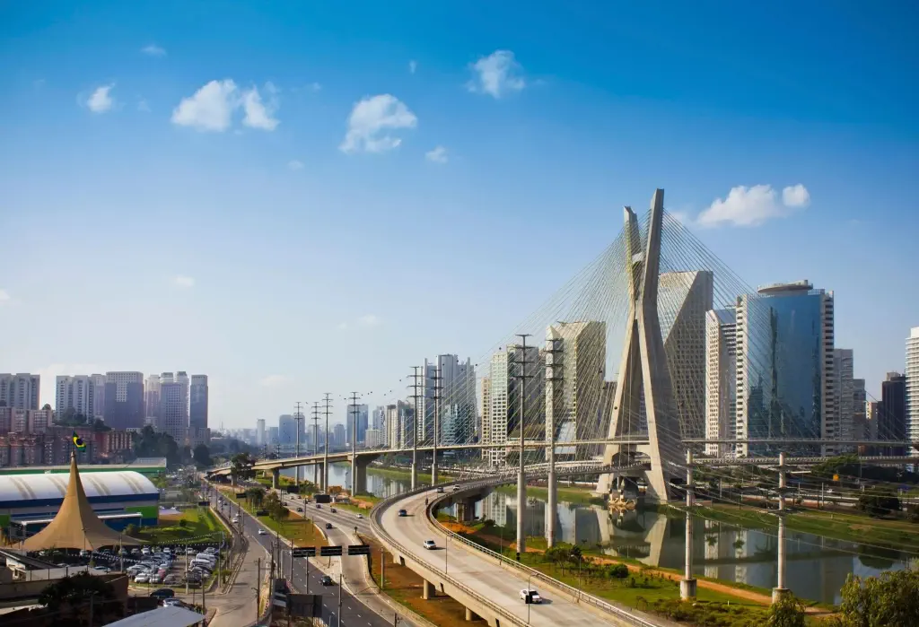 A cable-stayed bridge across a calm river bordered with roads and a cluster of high-rise buildings.