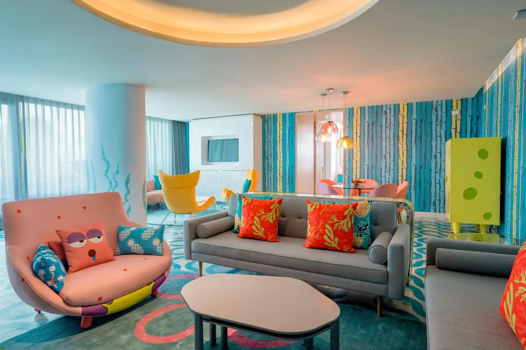 Colorful modern living room with a playful vibe. Features a gray sofa with vibrant pillows, a quirky face-patterned chair, and colorful walls with whimsical designs.