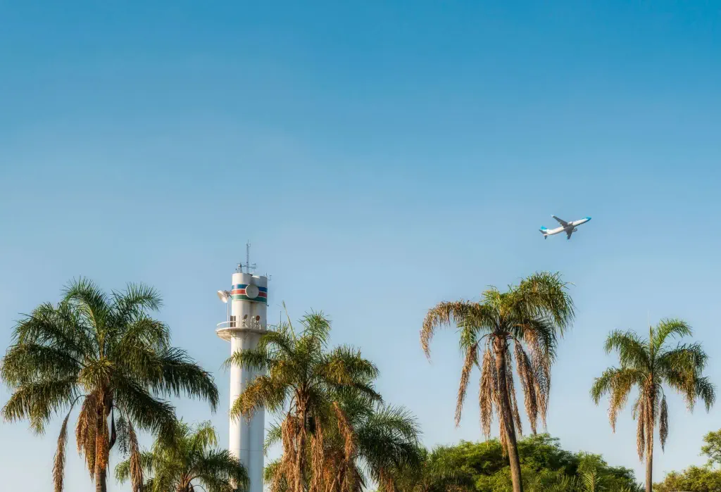 An airplane cruises the clear sky, framed by treelines and a prominent concrete tower in the foreground.