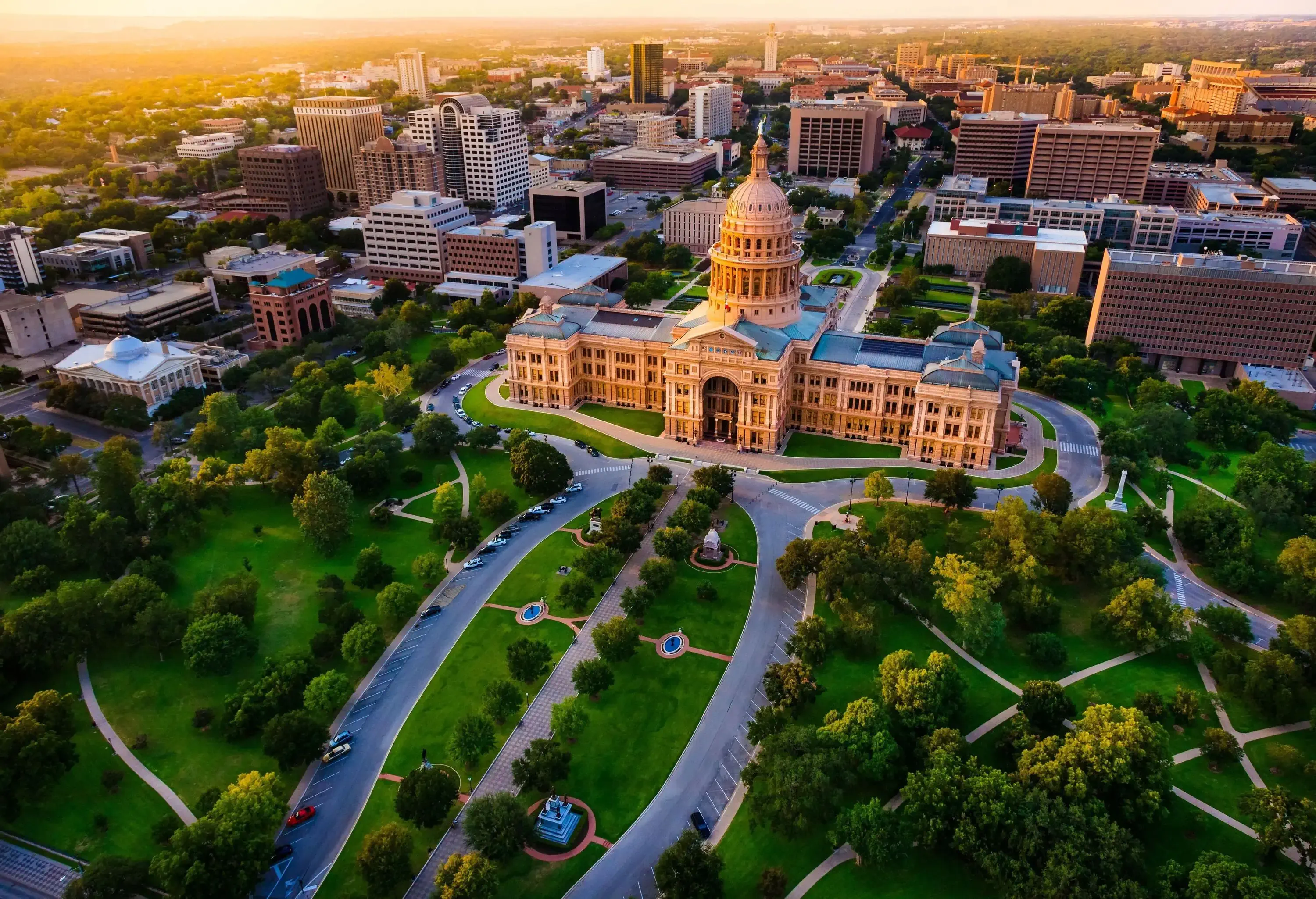 Texas State Capitol is a renaissance revival dome structure surrounded by a cluster of city buildings in front of a park with intersecting walkways across wide green lawns and lush trees.
