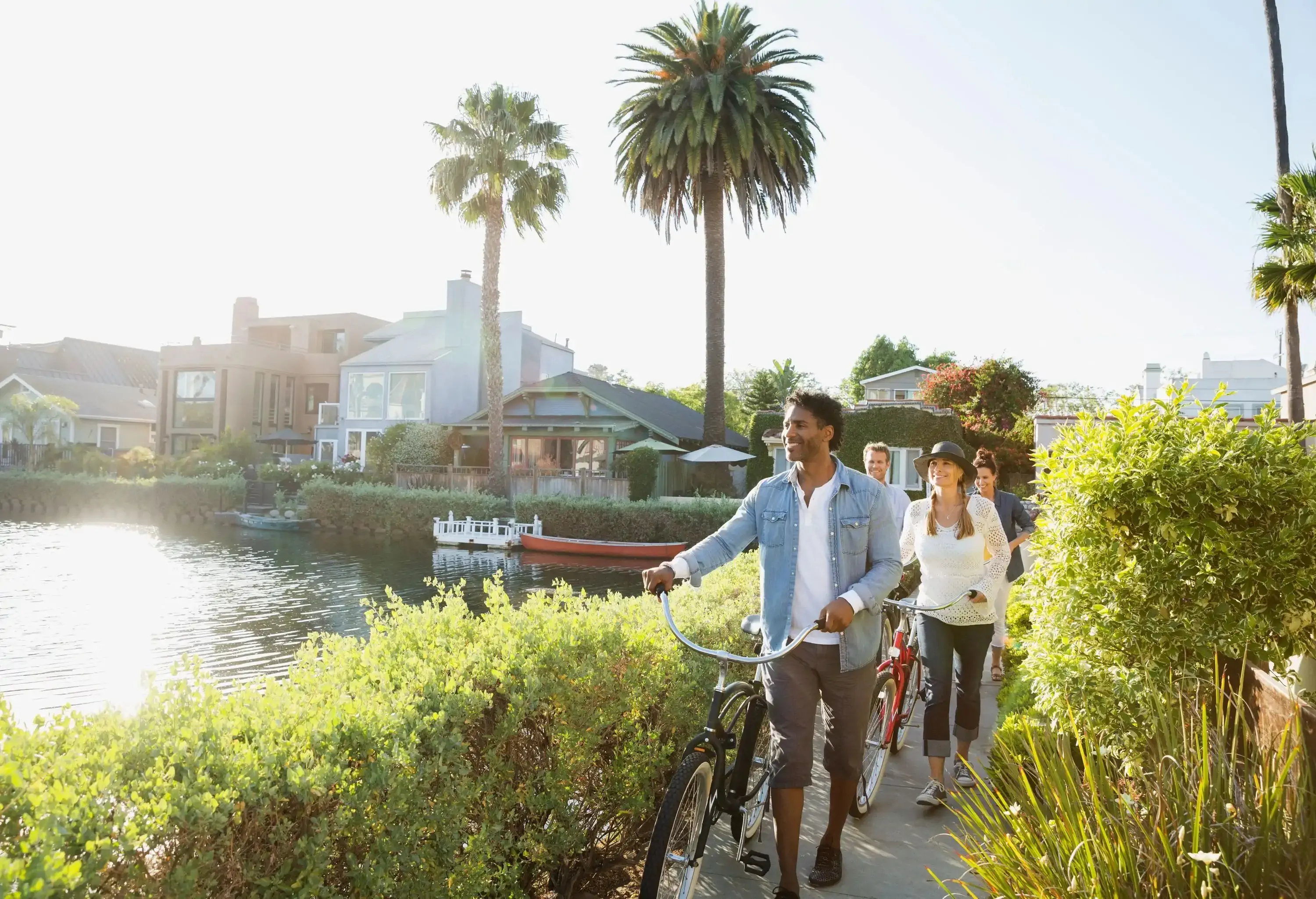 Four people walking their bikes on a narrow lane lined with bushes along a canal.