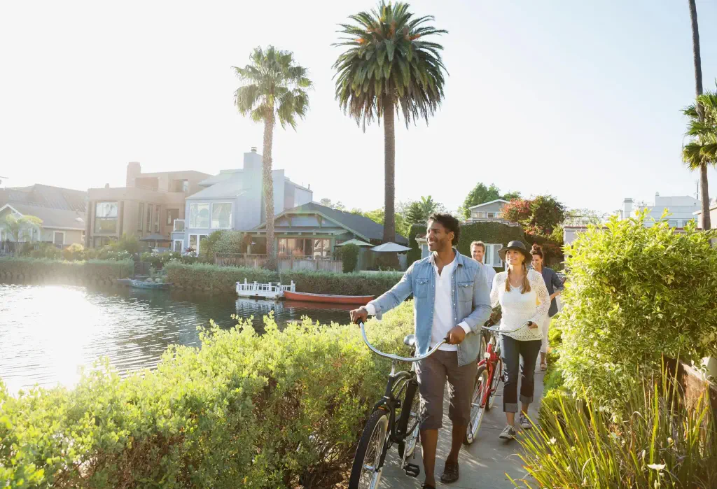 Four people walking their bikes on a narrow lane lined with bushes along a canal.