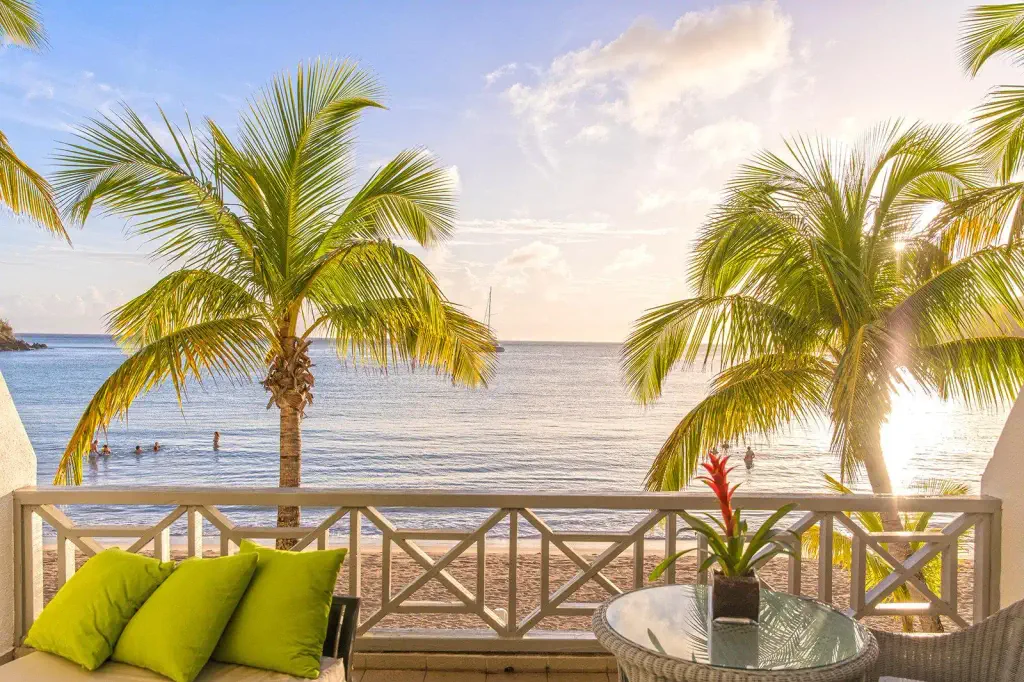 Balcony view of a serene beach at sunset with palm trees, soft waves, and people swimming. Cushioned seats and a vibrant plant enhance relaxation.