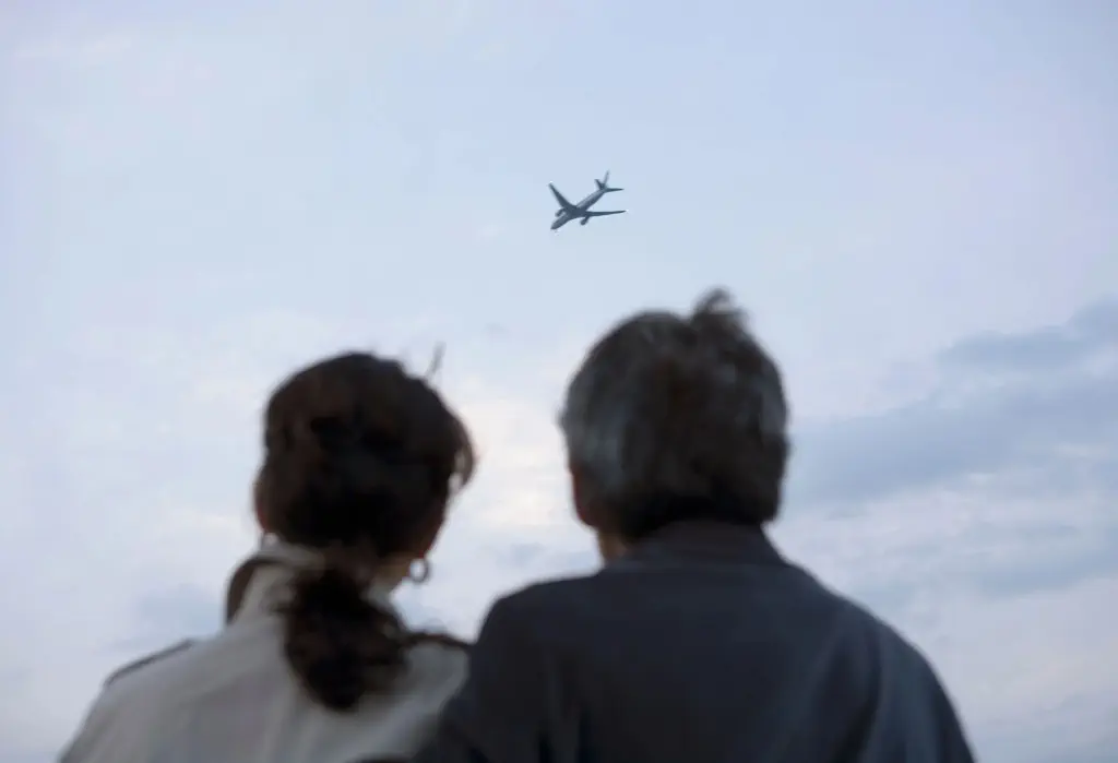 Back view of a couple standing side by side looks up at the airplane flying in the sky.