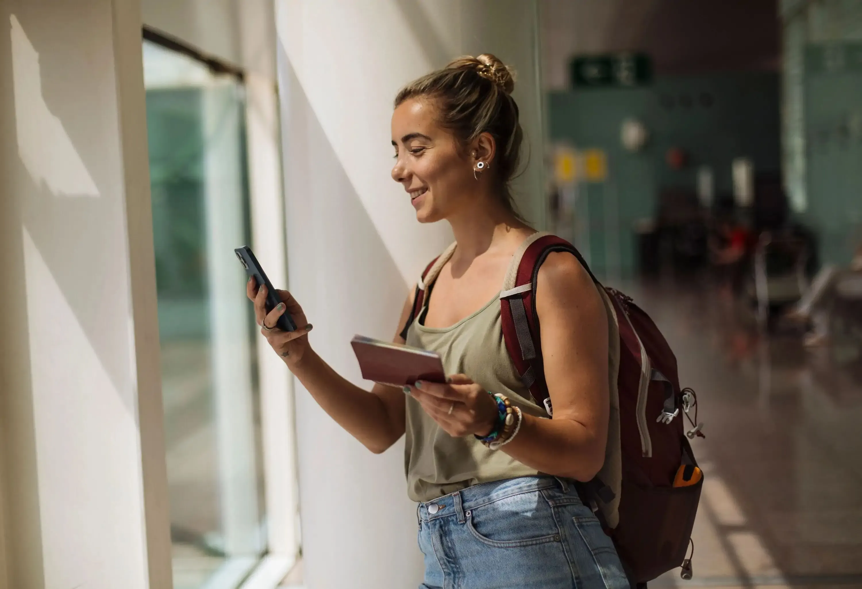 A young woman carrying a backpack and holding her passport is checking her phone.